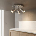 Chrome three-light adjustable ceiling spotlight illuminated above a modern white countertop in a minimalist kitchen.