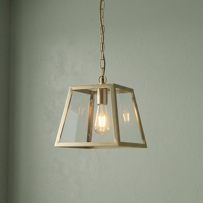 Antique brass pendant light with a tapered square glass shade hanging by a chain against a muted green wall.