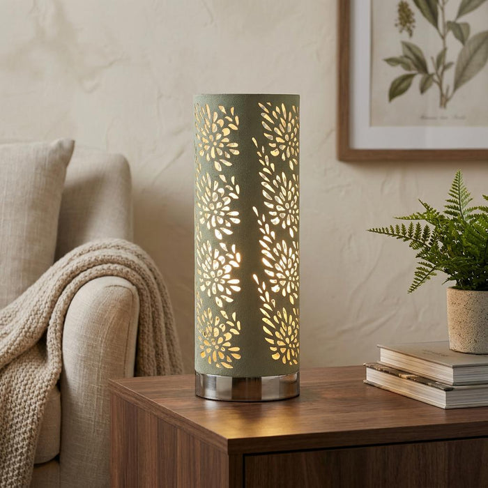 Sage green cylindrical table lamp with floral cutouts glowing on a wooden side table in a cozy living room.