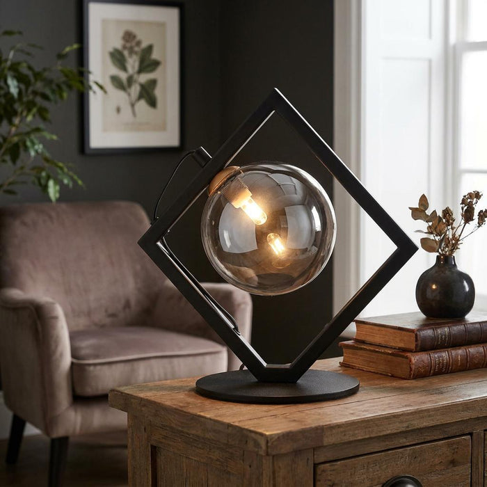 Modern table lamp featuring a black diamond-shaped frame and a glowing smoked glass sphere on a wooden table.
