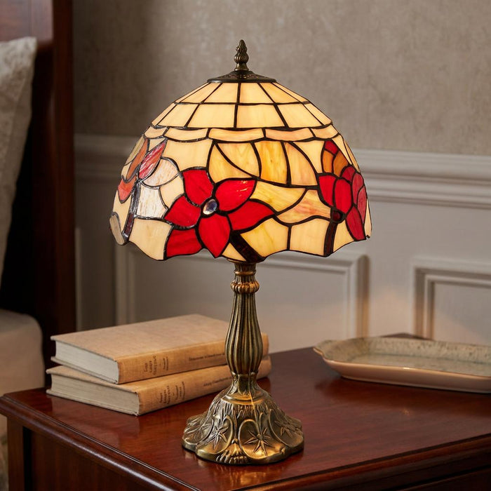Ornate Tiffany table lamp with red floral patterns on a cream glass shade, set on a dark wood bedside table with books.