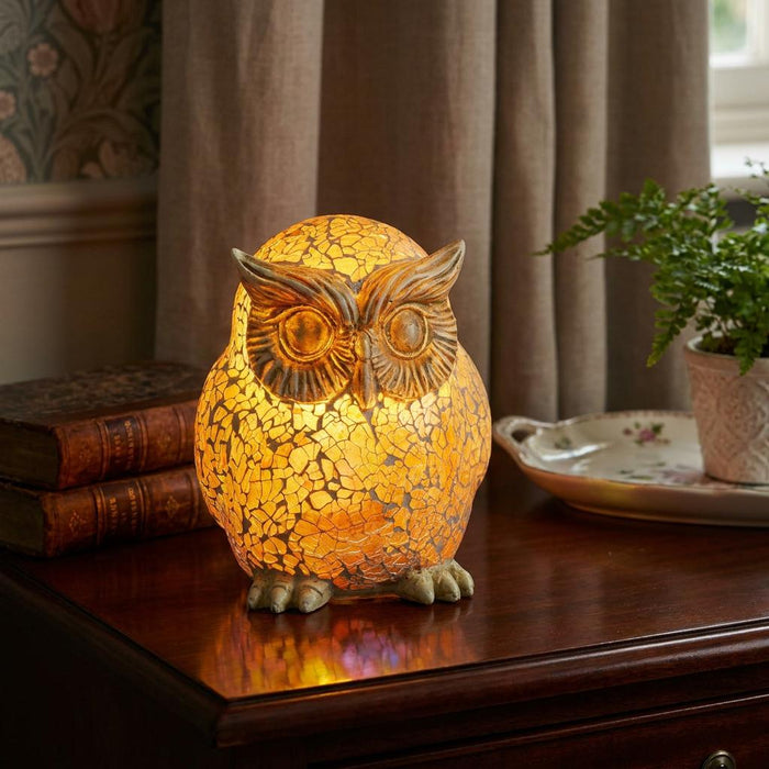 Warmly lit owl-shaped mosaic glass lamp on a dark wood table next to old books.