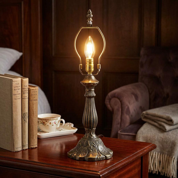 Ornate bronze-finished table lamp base with a lit candle bulb on a wooden bedside table near books and a tea set.