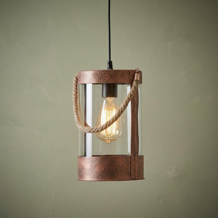 Cylindrical copper pendant light with glass shade, rope handle, and warm Edison bulb against a textured green wall.