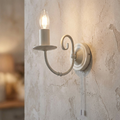Ivory candle-style wall light with a scroll arm and pull cord mounted on a textured, rustic plaster wall.