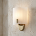 Single brass wall light with a curved, ribbed frosted glass shade glowing on a textured plaster wall.