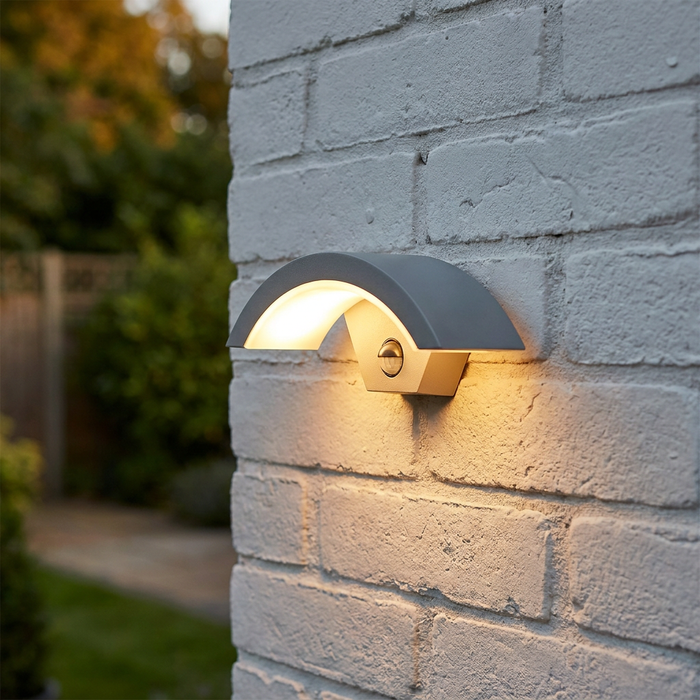 Silver grey arched outdoor LED wall light with a PIR sensor, illuminated on a white brick wall.