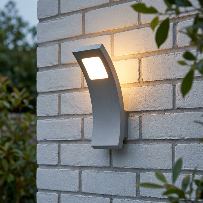 Curved silver-grey outdoor LED wall light emitting a warm glow on a white brick wall in a garden setting.