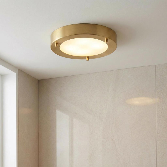 Round satin brass flush ceiling light with an opal glass diffuser, illuminated in a modern tiled bathroom.