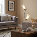 Two-light chrome table lamp with tulip-shaped shades on a wooden table in a modern living room setting.