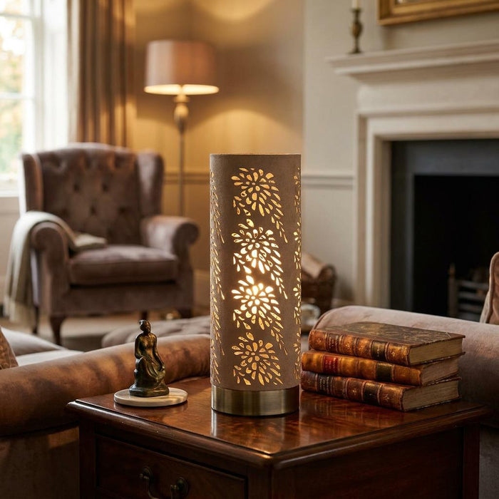 A cylindrical antique brass table lamp with floral cutouts glowing on a side table in a cozy living room.