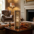 A cylindrical antique brass table lamp with floral cutouts glowing on a side table in a cozy living room.