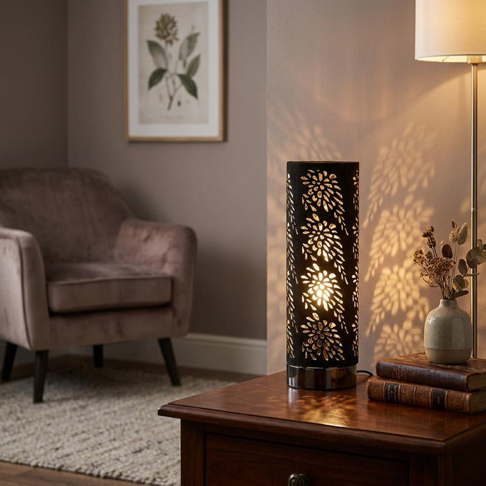 Black cylindrical table lamp with floral cutouts casting patterns on a wall in a living room.