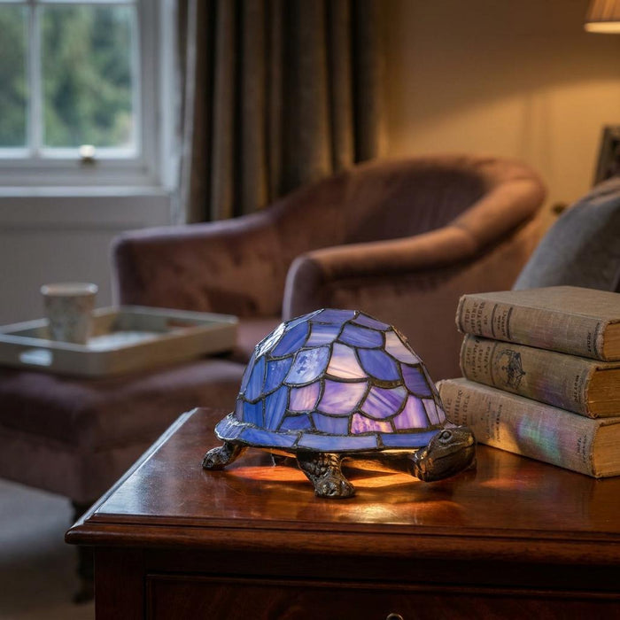 Tiffany-style tortoise lamp with blue and purple stained glass shell on a wooden table beside vintage books.