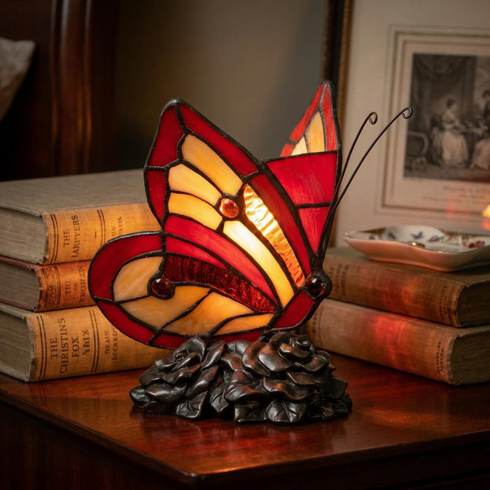 Glowing red and cream Tiffany butterfly lamp on a rose-shaped bronze base, set among vintage books on a wooden table.