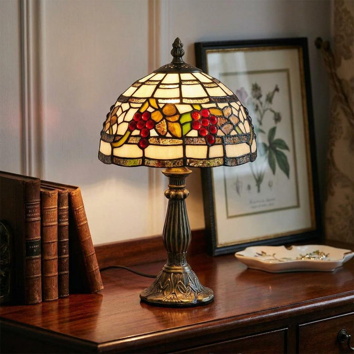 Stained glass Tiffany lamp with a red grape motif on a wooden desk next to vintage books and a framed picture.