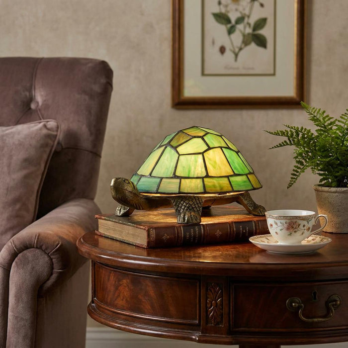 Green Tiffany-style tortoise lamp on a book beside a teacup on a side table.