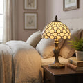 Tiffany-style lamp with a jewelled honey glass shade glowing on a dark wood bedside table in a bedroom.