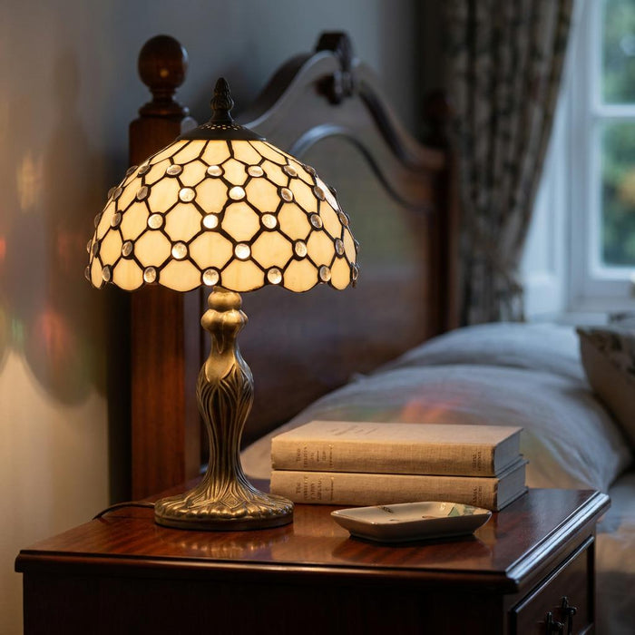 Lit Tiffany jewel table lamp with a scalloped cream shade on a wooden nightstand in a bedroom setting.