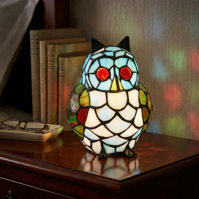 Stained glass owl table lamp with glowing red eyes on a wooden nightstand beside books.
