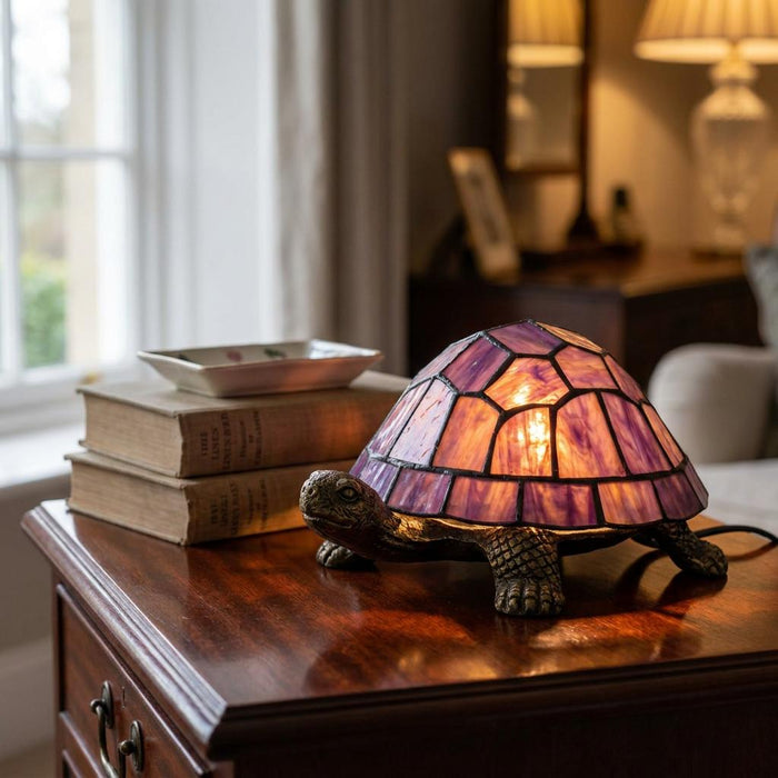Purple Tiffany-style tortoise table lamp glowing on a wooden chest next to a stack of books.