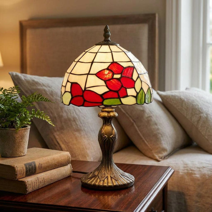 Tiffany-style lamp with a red floral stained glass shade on an ornate bronze base, set on a wooden bedside table.