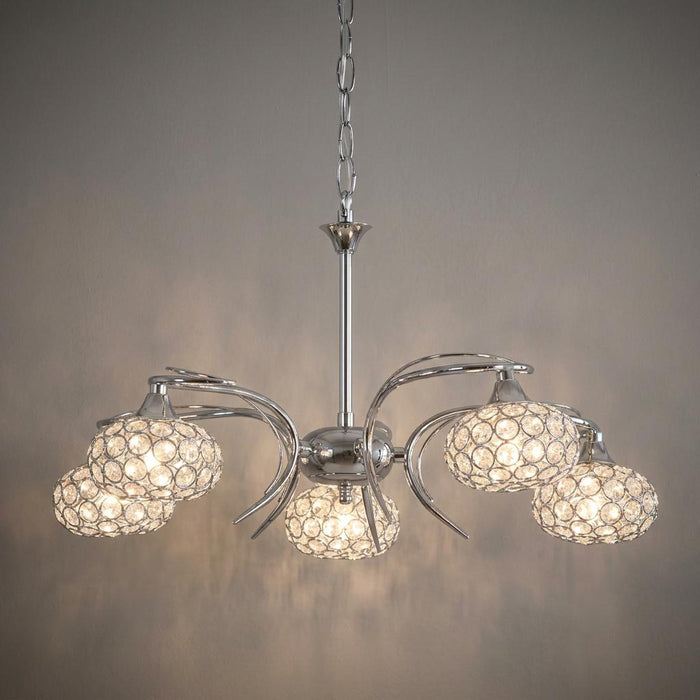 Five-light chrome chandelier with curved arms and sparkling crystal-beaded shades glowing against a neutral wall.