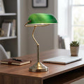 Antique brass bankers lamp with a glowing green glass shade sitting on a wooden desk in a modern home office.