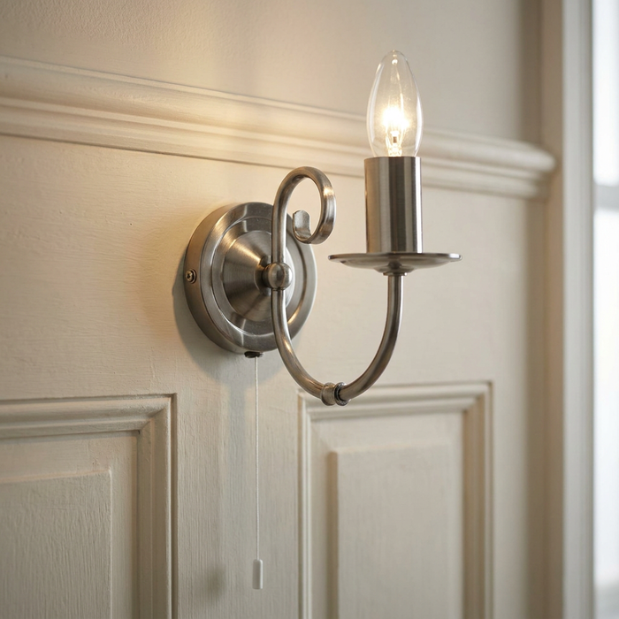 Antique silver wall light with a scroll arm and candle bulb, mounted on a cream paneled wall with a pull cord.