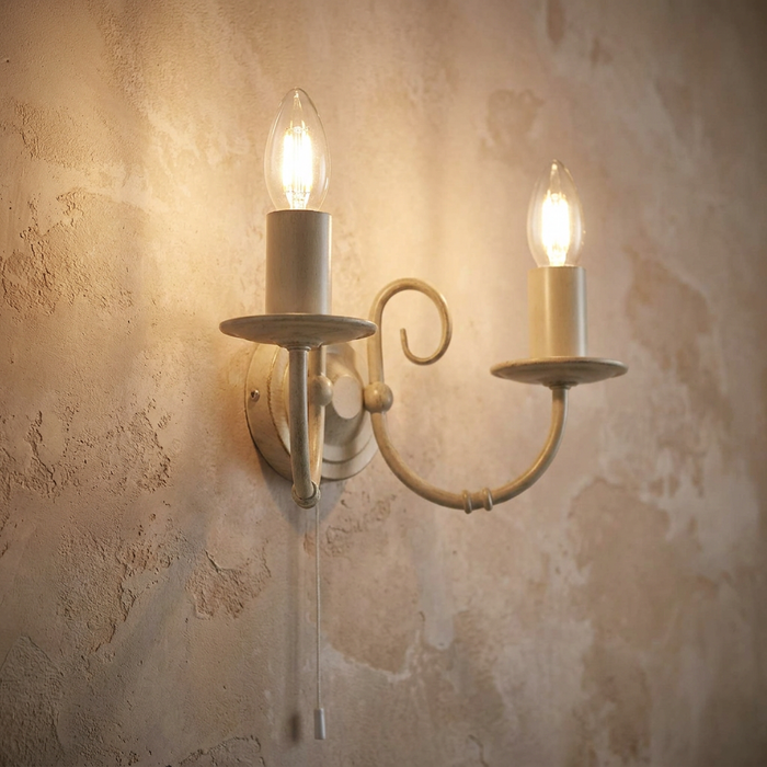 An ivory double wall light with two glowing candle bulbs mounted on a textured beige wall with a pull cord.