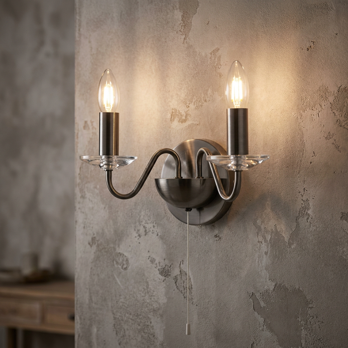 Two-light antique silver wall sconce with lit candle bulbs and glass accents on a distressed stone wall.