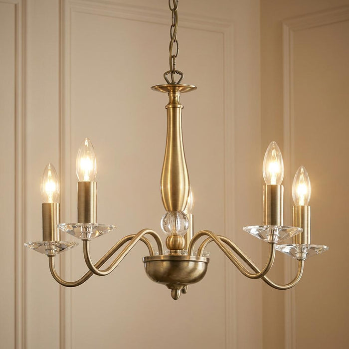 Five-light antique brass chandelier with curved arms, crystal drip pans, and candle-style bulbs in a neutral room.