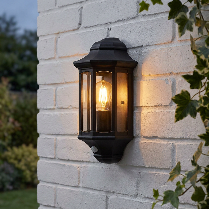 Black outdoor half-lantern wall light with PIR sensor and glowing filament bulb on a white brick wall.