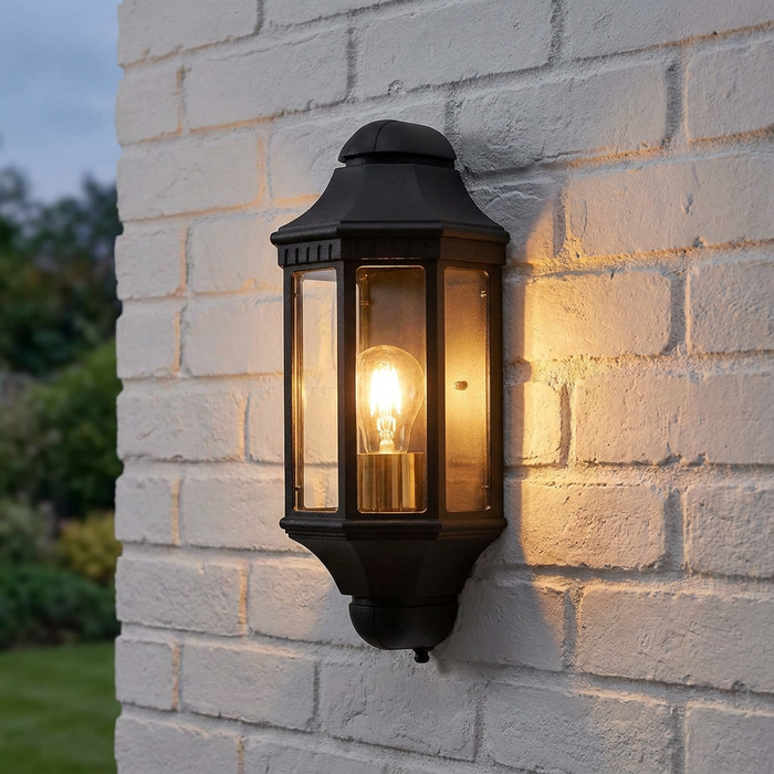 Traditional black outdoor wall lantern illuminated with warm light on a white brick wall.