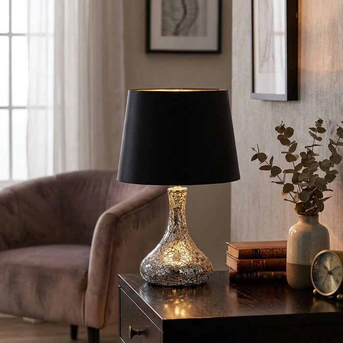Mirrored mosaic table lamp with a black shade sitting on a dark wood table in a warmly lit living room.