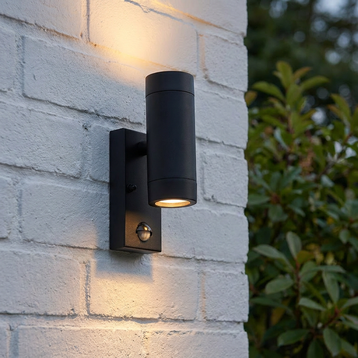 Matte black twin outdoor wall light with PIR sensor casting warm light on a white brick wall.