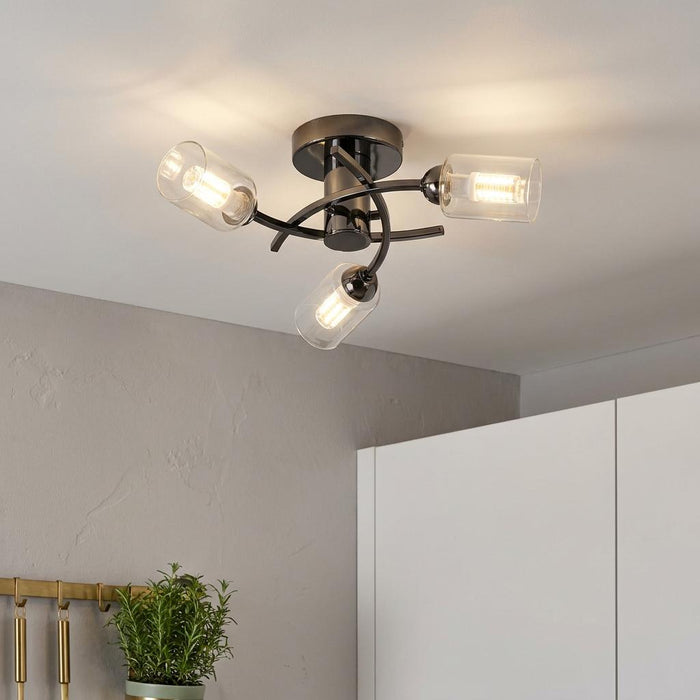 A black three-light flush ceiling fixture with curved arms and clear cylindrical glass shades lit in a kitchen.