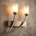 Oaks Ofira 2-light antique brass wall light with frosted glass shades on a textured wall.