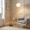 Otto Modern Matt Brass Arc Floor Lamp with a white globe shade next to a grey armchair in a neutral living room.