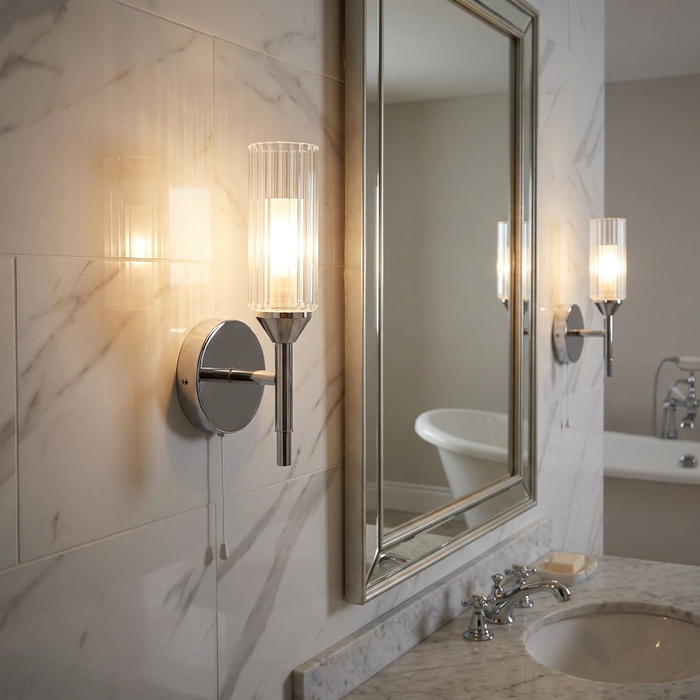 Chrome wall light with a fluted glass shade and pull cord mounted on a marble-tiled bathroom wall.