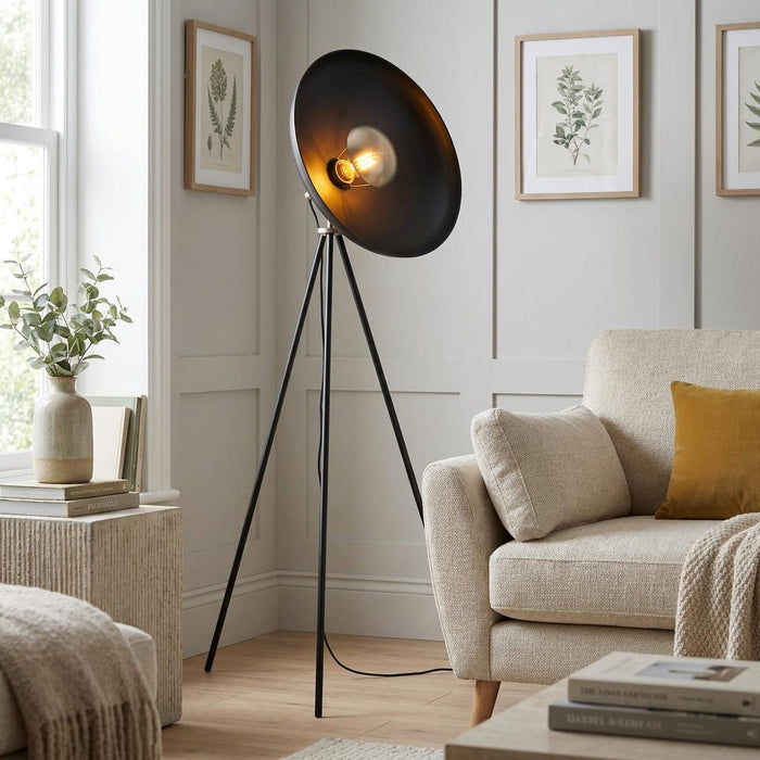 An Ovni black industrial tripod floor lamp with a large dish shade illuminates a cozy living room corner.