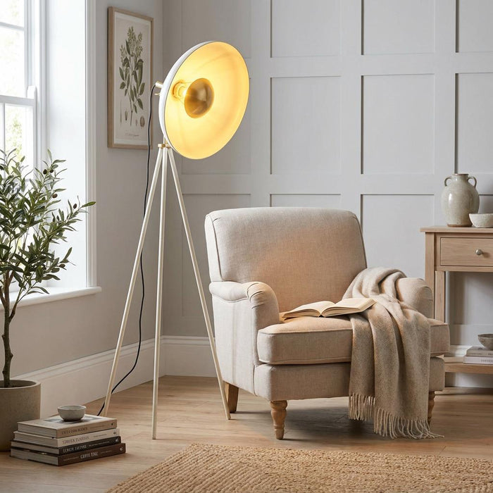 Ovni white industrial tripod floor lamp with a gold-lined dish shade next to a beige armchair in a living room.