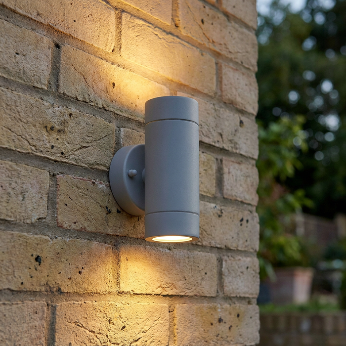 Grey cylindrical up-and-down outdoor wall light mounted on a brick wall and illuminated.