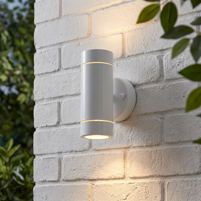 Sleek white cylindrical up and down outdoor wall light mounted and illuminated on a white brick wall.