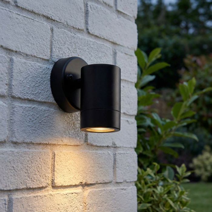 Palin black outdoor wall light casting a warm downward glow on a white brick wall in a garden setting.