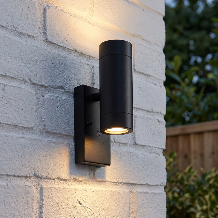 Modern black cylindrical up-and-down wall light mounted on a white brick wall, casting warm light.