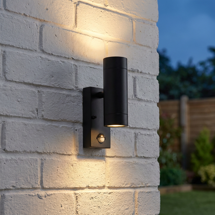 Black up and down outdoor wall light with a PIR sensor mounted on a white brick wall, emitting warm light.