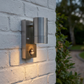 Brushed stainless steel outdoor wall downlight with a PIR sensor mounted on a white brick wall.