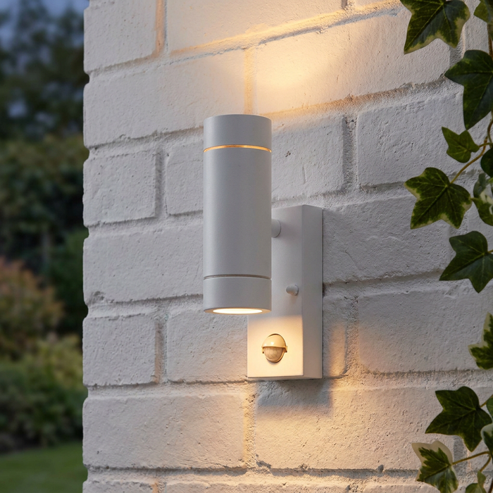 White up and down outdoor wall light with PIR sensor, casting warm light onto a white brick wall.