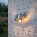 Brushed stainless steel twin outdoor wall spotlights illuminated on a white brick wall.